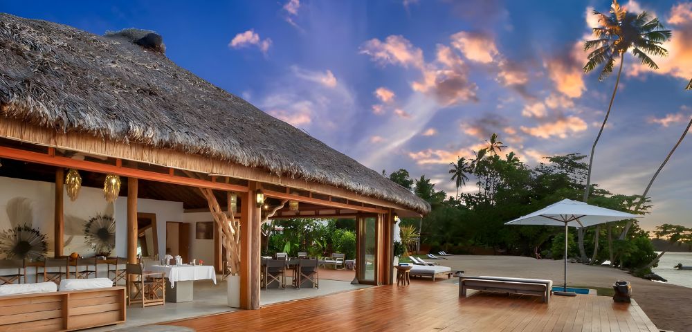 Tropical villa with a thatched roof, wooden deck, dining area, and beach view under a colorful sunset sky, surrounded by palm trees.