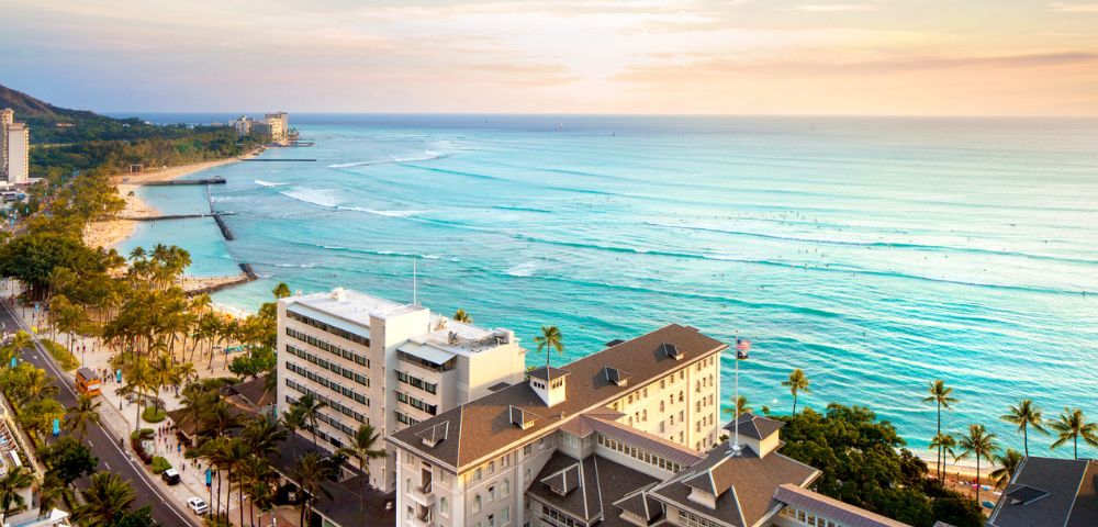 Aerial view of Waikiki Beach, showcasing turquoise waves, surfers, palm trees, and hotels under a colorful sunset sky.