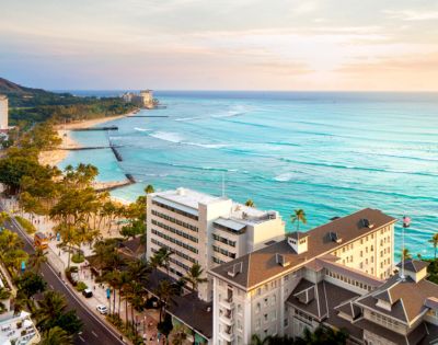 Aerial view of Waikiki Beach, showcasing turquoise waves, surfers, palm trees, and hotels under a colorful sunset sky.