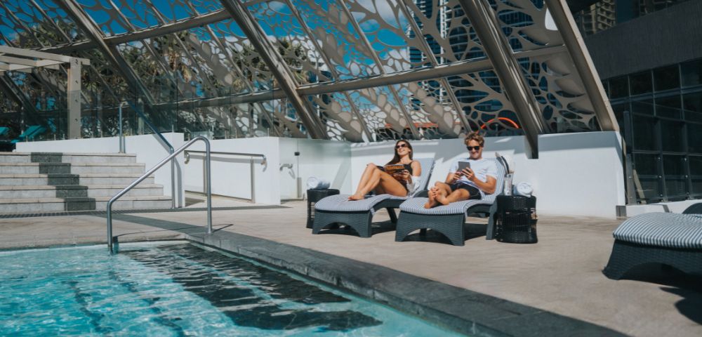 A woman in a bikini and a man in swimwear relax on lounge chairs by a pool under a glass roof, enjoying the sunny day.