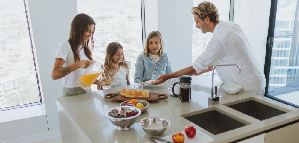 A family of four enjoying breakfast at a kitchen island in a modern, well-lit kitchen. A woman pours orange juice, two young girls look on, and a man serves a plate of food. The island features a wooden cutting board with bread, fruit in bowls, and a coffee maker.