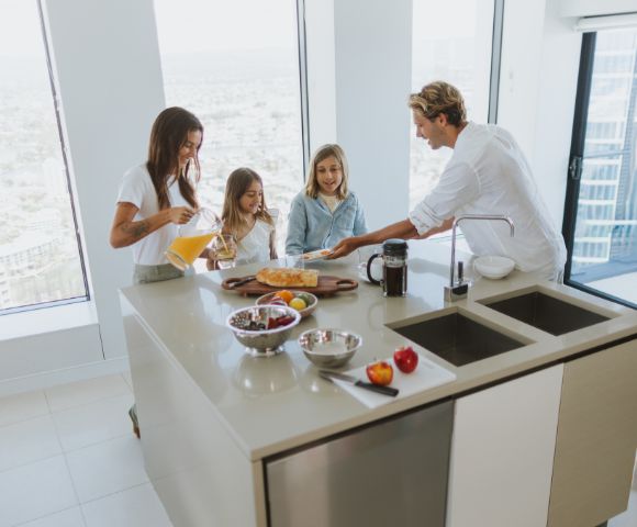 A family of four enjoying breakfast at a kitchen island in a modern, well-lit kitchen. A woman pours orange juice, two young girls look on, and a man serves a plate of food. The island features a wooden cutting board with bread, fruit in bowls, and a coffee maker.