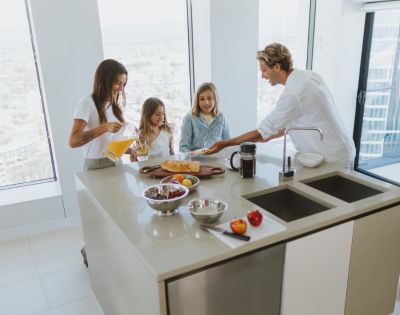 A family of four enjoying breakfast at a kitchen island in a modern, well-lit kitchen. A woman pours orange juice, two young girls look on, and a man serves a plate of food. The island features a wooden cutting board with bread, fruit in bowls, and a coffee maker.