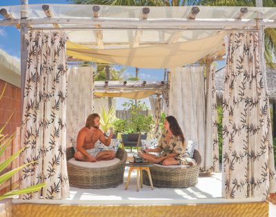 A couple relaxing in a shaded outdoor cabana with seating and curtains, enjoying drinks, with tropical foliage and blue sky visible in the background.