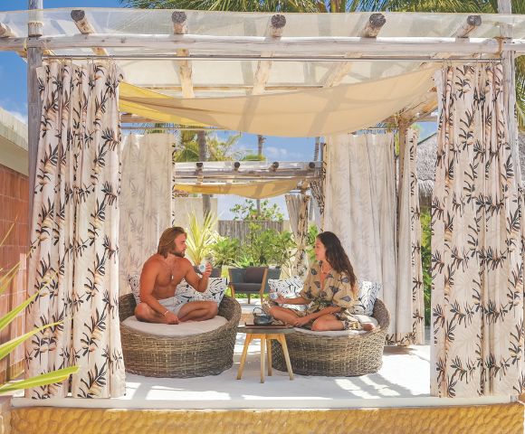 A couple relaxing in a shaded outdoor cabana with seating and curtains, enjoying drinks, with tropical foliage and blue sky visible in the background.