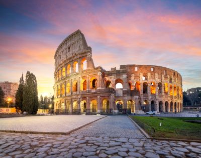 The photo depicts the Colosseum in Rome, illuminated by warm lights against a vibrant sunset sky, surrounded by cobblestone pathways and lush greenery.