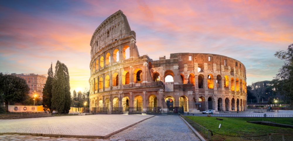 The photo depicts the Colosseum in Rome, illuminated by warm lights against a vibrant sunset sky, surrounded by cobblestone pathways and lush greenery.