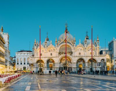 The image depicts the stunning exterior of St. Mark's Basilica in Venice, featuring its intricate architecture, domes, and decorative details, with empty outdoor dining tables arranged in the foreground against a twilight sky.