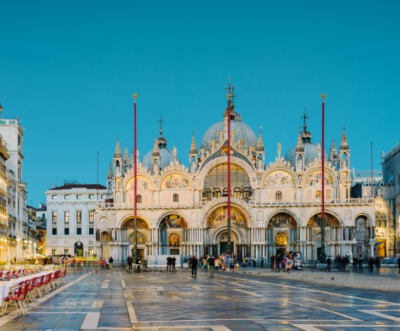 The image depicts the stunning exterior of St. Mark's Basilica in Venice, featuring its intricate architecture, domes, and decorative details, with empty outdoor dining tables arranged in the foreground against a twilight sky.