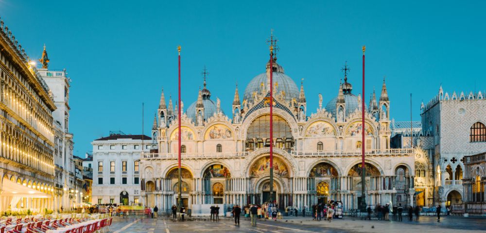 The image depicts the stunning exterior of St. Mark's Basilica in Venice, featuring its intricate architecture, domes, and decorative details, with empty outdoor dining tables arranged in the foreground against a twilight sky.