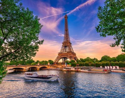 The image features the iconic Eiffel Tower in Paris, framed by lush green trees, with a boat sailing on the Seine River amidst a sunset sky adorned with wispy clouds.