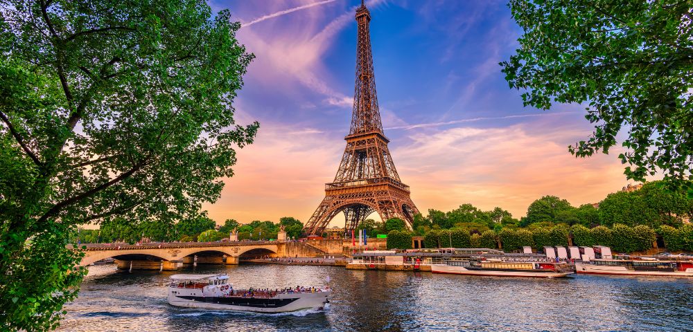 The image features the iconic Eiffel Tower in Paris, framed by lush green trees, with a boat sailing on the Seine River amidst a sunset sky adorned with wispy clouds.