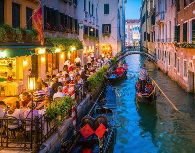 A charming Venetian canal scene with gondolas, outdoor dining, and illuminated buildings reflecting the evening's vibrant atmosphere.