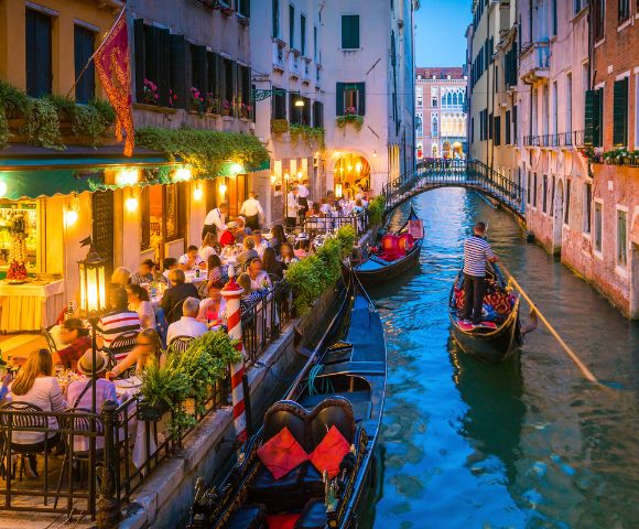 A charming Venetian canal scene with gondolas, outdoor dining, and illuminated buildings reflecting the evening's vibrant atmosphere.