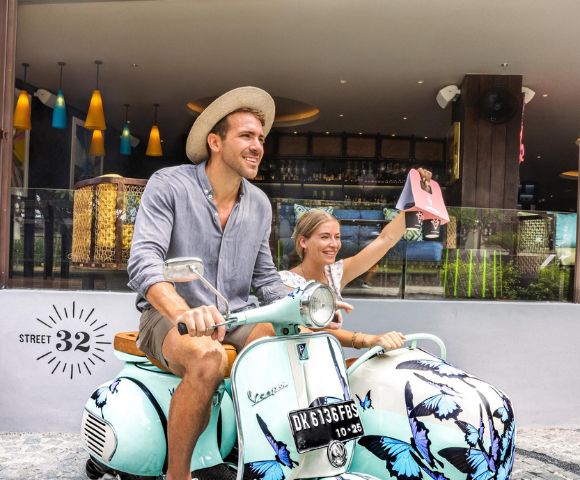 A couple enjoys a playful moment on a vintage scooter adorned with butterfly designs, set against a vibrant café backdrop.