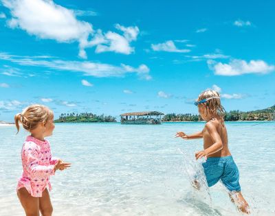 Two children play joyfully in shallow turquoise waters under a bright blue sky, with a distant boat and lush greenery in the background.