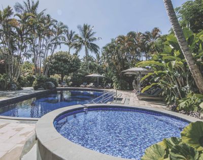Tropical pool area featuring a large blue swimming pool, a smaller hot tub, lush palm trees, and sun loungers under umbrellas.