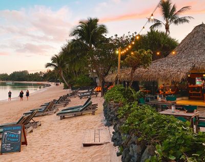 Tropical beach at sunset with palm trees, a thatched-roof café, and lounge chairs along the sand. Warm lighting and a tranquil, inviting atmosphere.