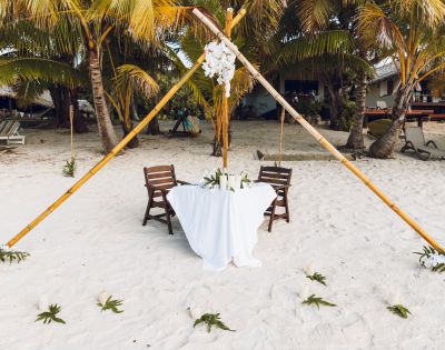 A romantic beach setting features a small table with a white cloth and two chairs under a bamboo canopy adorned with white flowers, surrounded by palm trees.