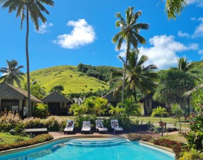 Tropical resort scene with a clear pool, lined with white loungers and towels. Thatched huts are surrounded by lush greenery and tall palm trees under a bright blue sky.
