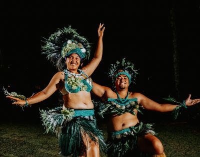 Two dancers in vibrant Polynesian attire perform under a dark sky. Feathers and grass skirts add energy and cultural flair. They're smiling and expressive.