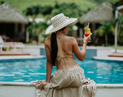 Woman in a straw hat sits by a pool, wearing a bikini and a flowing skirt. She holds a tropical drink, exuding a relaxed, vacation vibe.