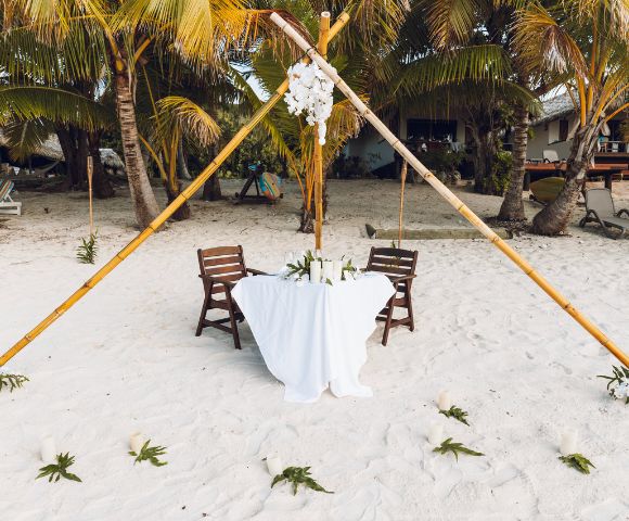 A romantic beach setting features a small table with a white cloth and two chairs under a bamboo canopy adorned with white flowers, surrounded by palm trees.