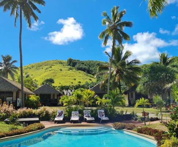 Tropical resort scene with a clear pool, lined with white loungers and towels. Thatched huts are surrounded by lush greenery and tall palm trees under a bright blue sky.