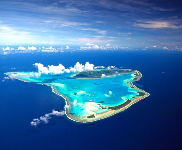 Aerial view of a heart-shaped island surrounded by vibrant turquoise waters, lush greenery, and a clear blue sky with scattered clouds above.