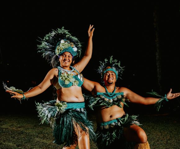 Two dancers in vibrant Polynesian attire perform under a dark sky. Feathers and grass skirts add energy and cultural flair. They're smiling and expressive.