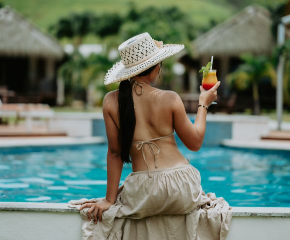 Woman in a straw hat sits by a pool, wearing a bikini and a flowing skirt. She holds a tropical drink, exuding a relaxed, vacation vibe.
