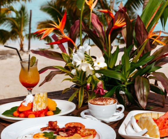 Tropical brunch setup on a beach with vibrant flowers. A fruity cocktail and a plate with fruit slices sit beside a cappuccino. Relaxing, exotic vibe.