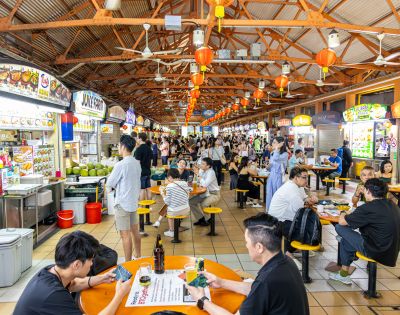 Bustling hawker center with people dining at orange tables under a wooden roof. Bright food stalls line the sides, creating a lively atmosphere.