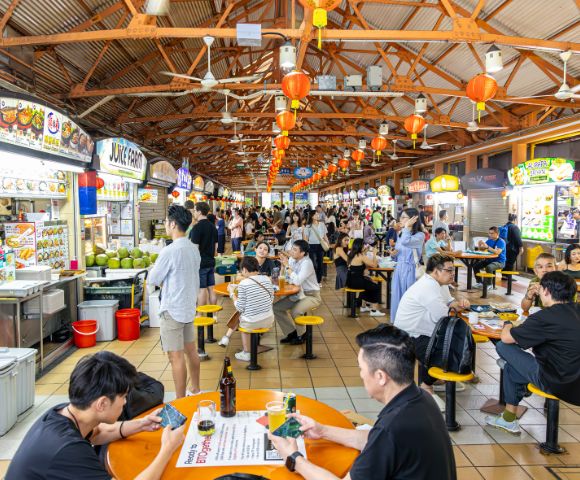 Bustling hawker center with people dining at orange tables under a wooden roof. Bright food stalls line the sides, creating a lively atmosphere.