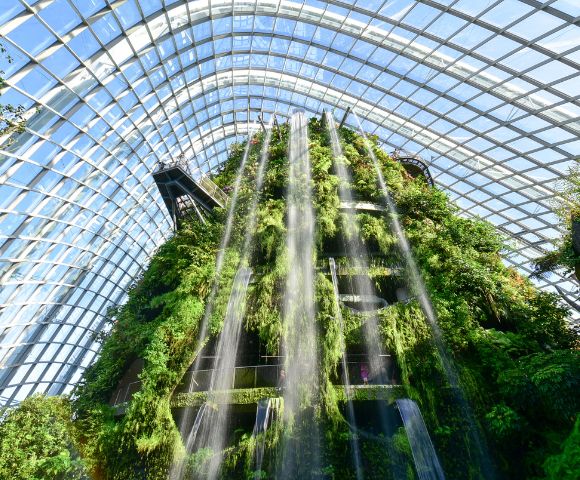 Alt text: Lush indoor waterfall cascades down a vertical garden within a sunlit glass dome, creating a refreshing, natural oasis atmosphere.