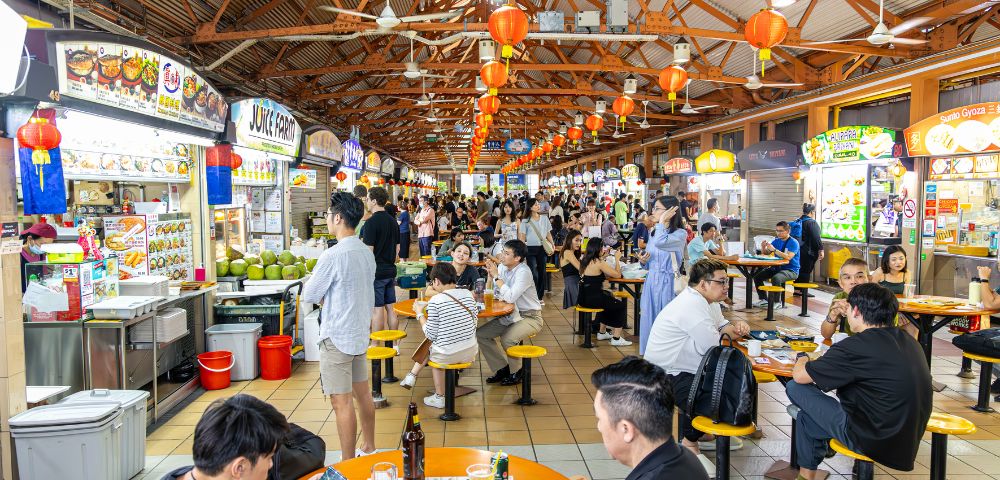 Bustling hawker center with people dining at orange tables under a wooden roof. Bright food stalls line the sides, creating a lively atmosphere.