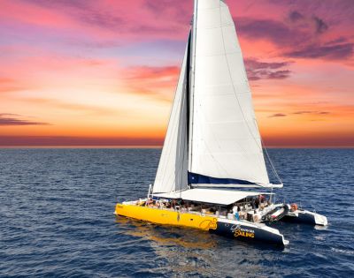 A large yellow catamaran with white sails glides across the ocean at sunset, under a breathtaking sky painted in vibrant pink, orange, and purple hues.
