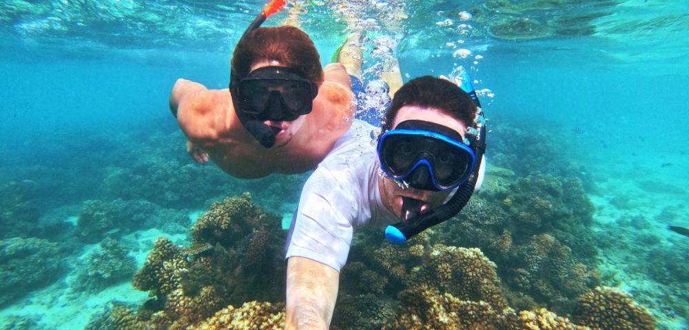 Two people snorkel in clear turquoise water above vibrant coral reefs. They wear snorkels and masks, capturing a joyful underwater adventure selfie.