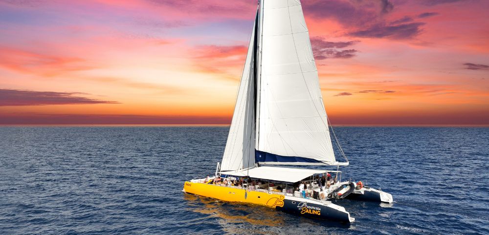 A large yellow catamaran with white sails glides across the ocean at sunset, under a breathtaking sky painted in vibrant pink, orange, and purple hues.