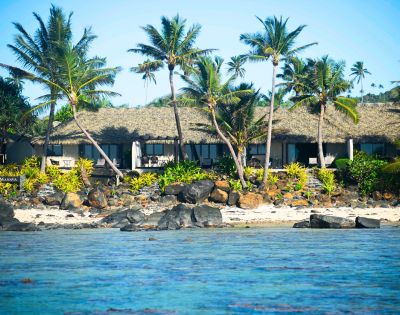 Tropical beach scene featuring a thatched-roof resort surrounded by palm trees, rocky shore, and vibrant greenery by the water's edge.
