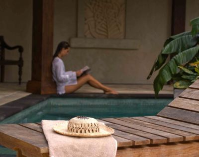 A cozy outdoor setting near a pool, featuring a straw hat on a wooden lounge chair and a person reading in the background.