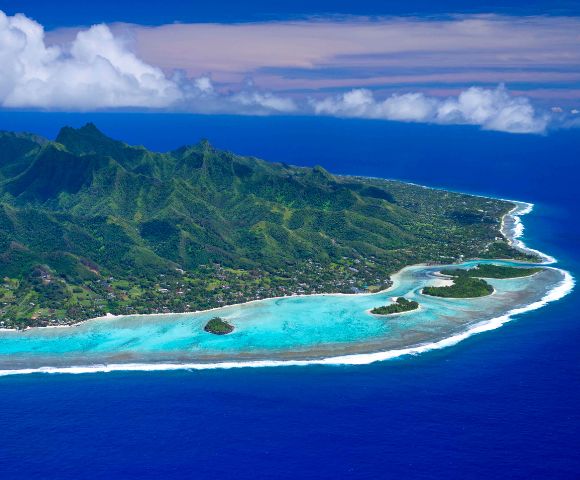 Aerial view of a lush, green island with turquoise waters, white sandy beaches, and scattered rocky islets against a blue sky.