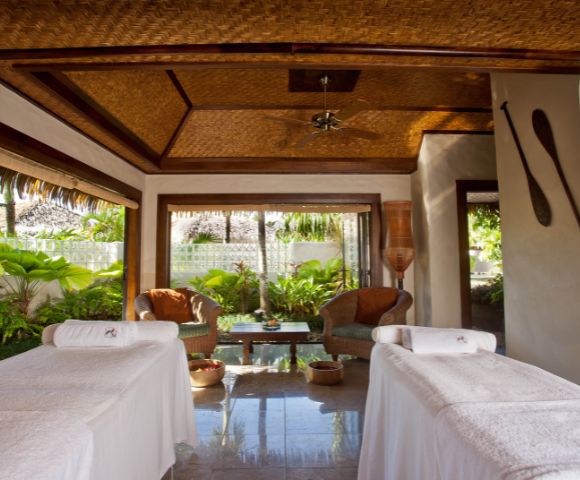 Luxury spa room with two massage tables draped with white towels, facing each other with a view of lush tropical greenery through an open window; wicker chairs and a small table with flowers are between the tables, and woven ceiling panels with a ceiling fan above.