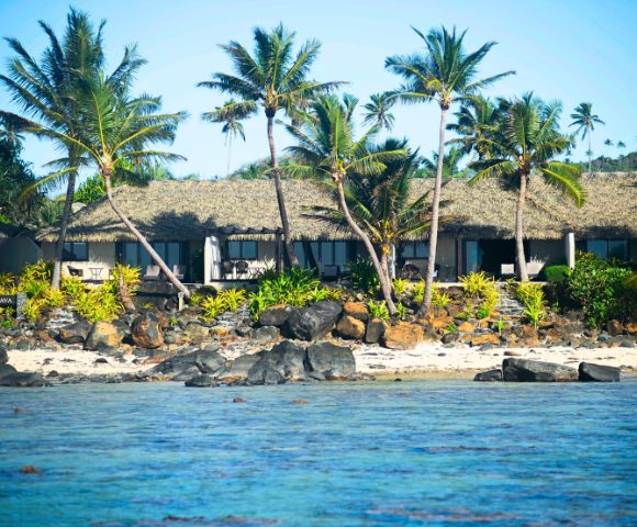 Tropical beach scene featuring a thatched-roof resort surrounded by palm trees, rocky shore, and vibrant greenery by the water's edge.