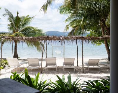Tranquil beach scene with five lounge chairs under a rustic canopy, surrounded by palm trees. Gentle waves and distant mountains create a serene atmosphere.