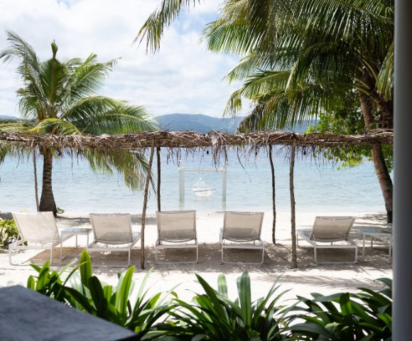 Tranquil beach scene with five lounge chairs under a rustic canopy, surrounded by palm trees. Gentle waves and distant mountains create a serene atmosphere.
