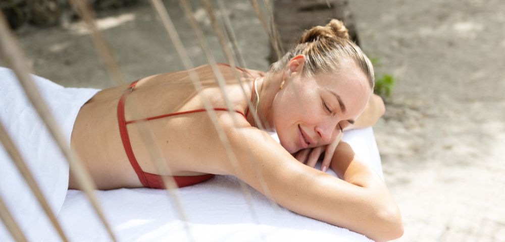 Woman in a red bikini relaxes on a white towel outdoors, eyes closed, smiling gently, with a serene and tranquil beach background.