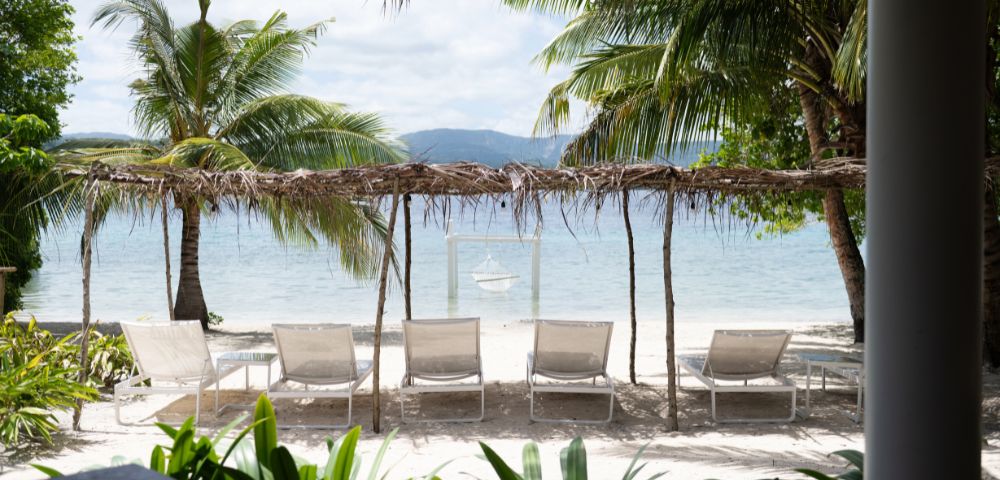 Tranquil beach scene with five lounge chairs under a rustic canopy, surrounded by palm trees. Gentle waves and distant mountains create a serene atmosphere.