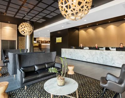 Modern hotel lobby with dark sofas, patterned carpet, and wooden stools. Three people sit at a marble reception desk. Warm lighting with a calm ambiance.
