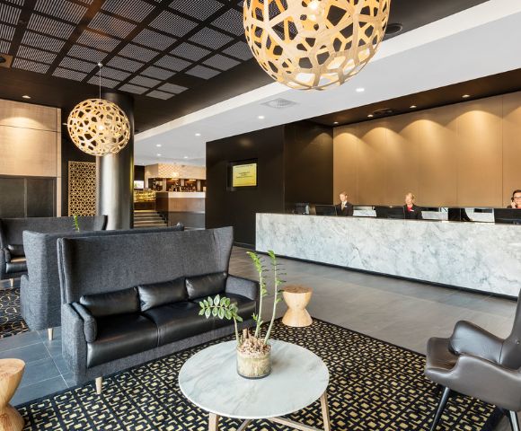 Modern hotel lobby with dark sofas, patterned carpet, and wooden stools. Three people sit at a marble reception desk. Warm lighting with a calm ambiance.